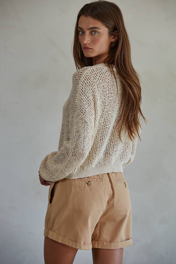 Model wearing a loose-knit cream Wren Sweater with button detail, paired with tan shorts.