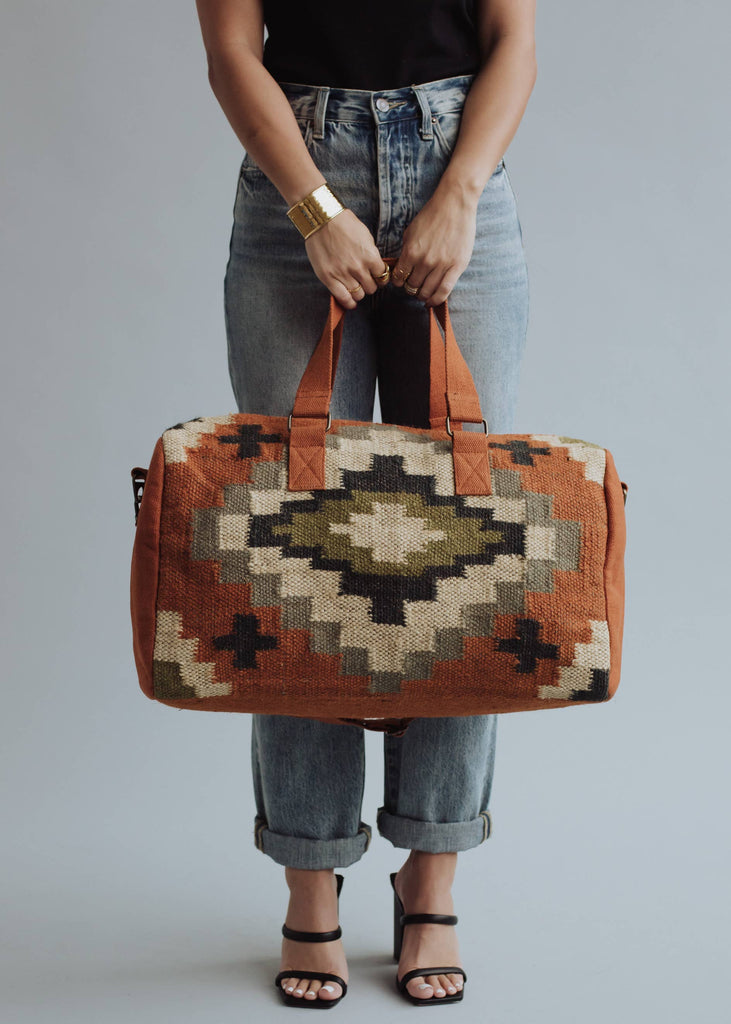 Earth-tone woven weekender bag with geometric pattern in black, green, and cream, held by woman in jeans and black top
