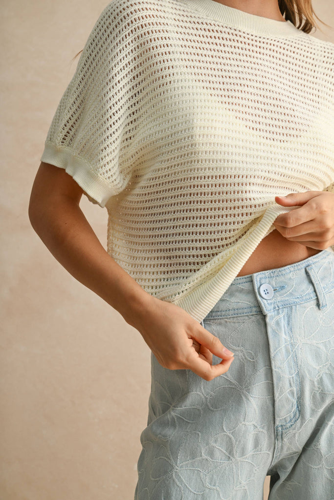 Model wearing a cream open-knit short sleeve sweater with open back detail, paired with embroidered light-wash denim pants.