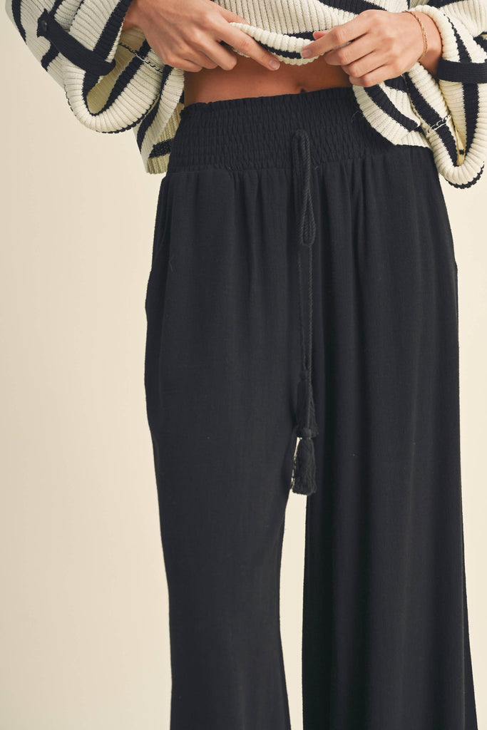 Woman wearing black wide-leg pants with smocked waistband and tassel drawstring.