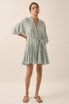 Woman wearing a sage green button-down mini dress with a braided waist tie and flowy hemline.