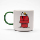 White ceramic mug with a green handle, featuring a beagle and a bird on a red doghouse, along with a group of characters enjoying music.