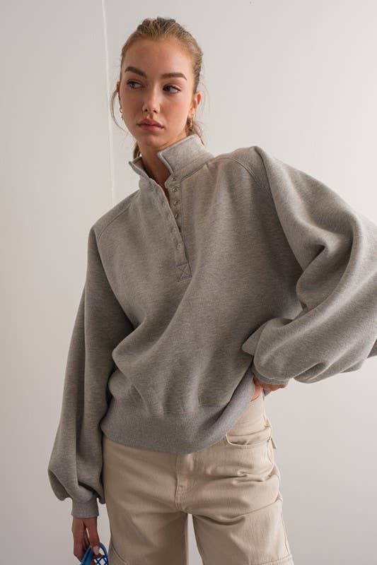 Woman wearing an oversized gray pullover sweatshirt with quarter snaps and voluminous sleeves, paired with beige pants.