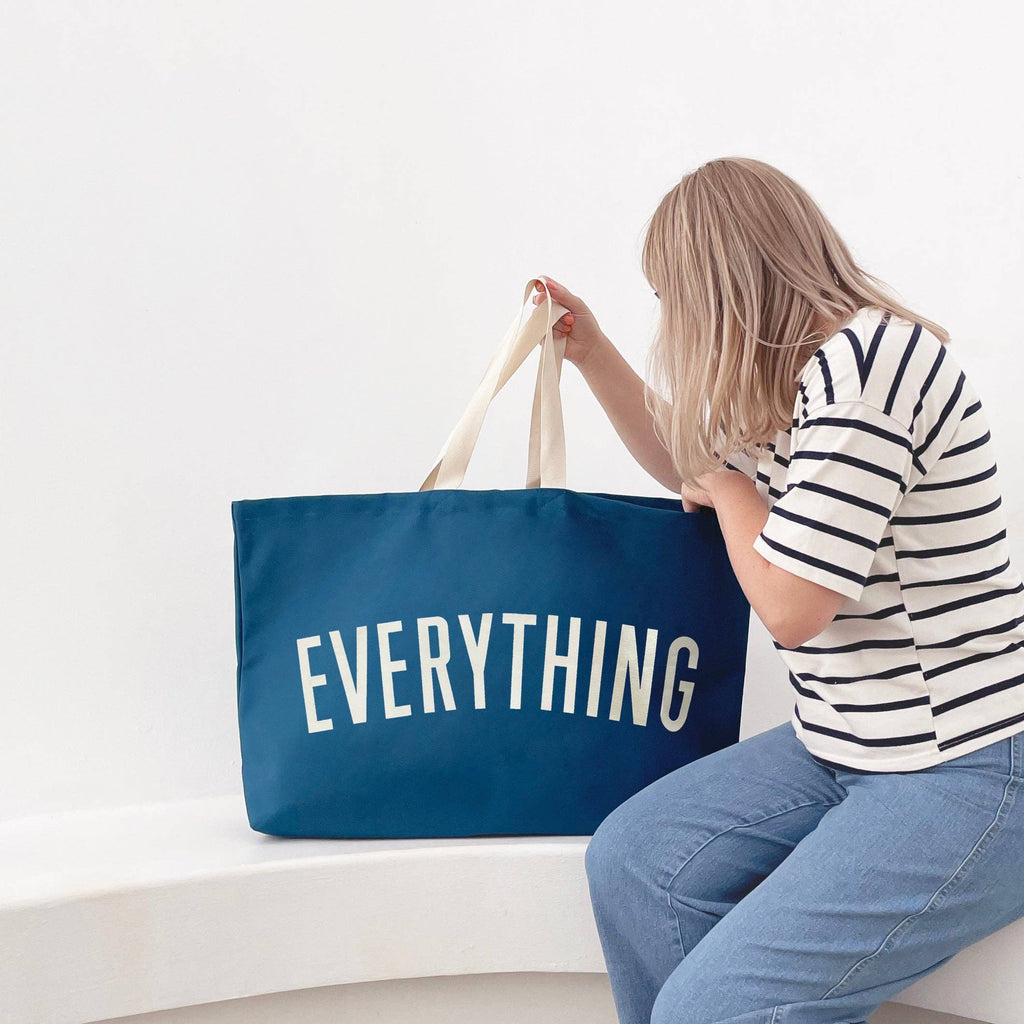 Oversized navy blue tote bag with cream handles and bold white “EVERYTHING” text, held by a hand against a light wood background