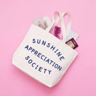 Cream canvas tote bag with “Sunshine Appreciation Society” printed in navy, filled with flip-flops, sunglasses, and travel essentials on a pink background