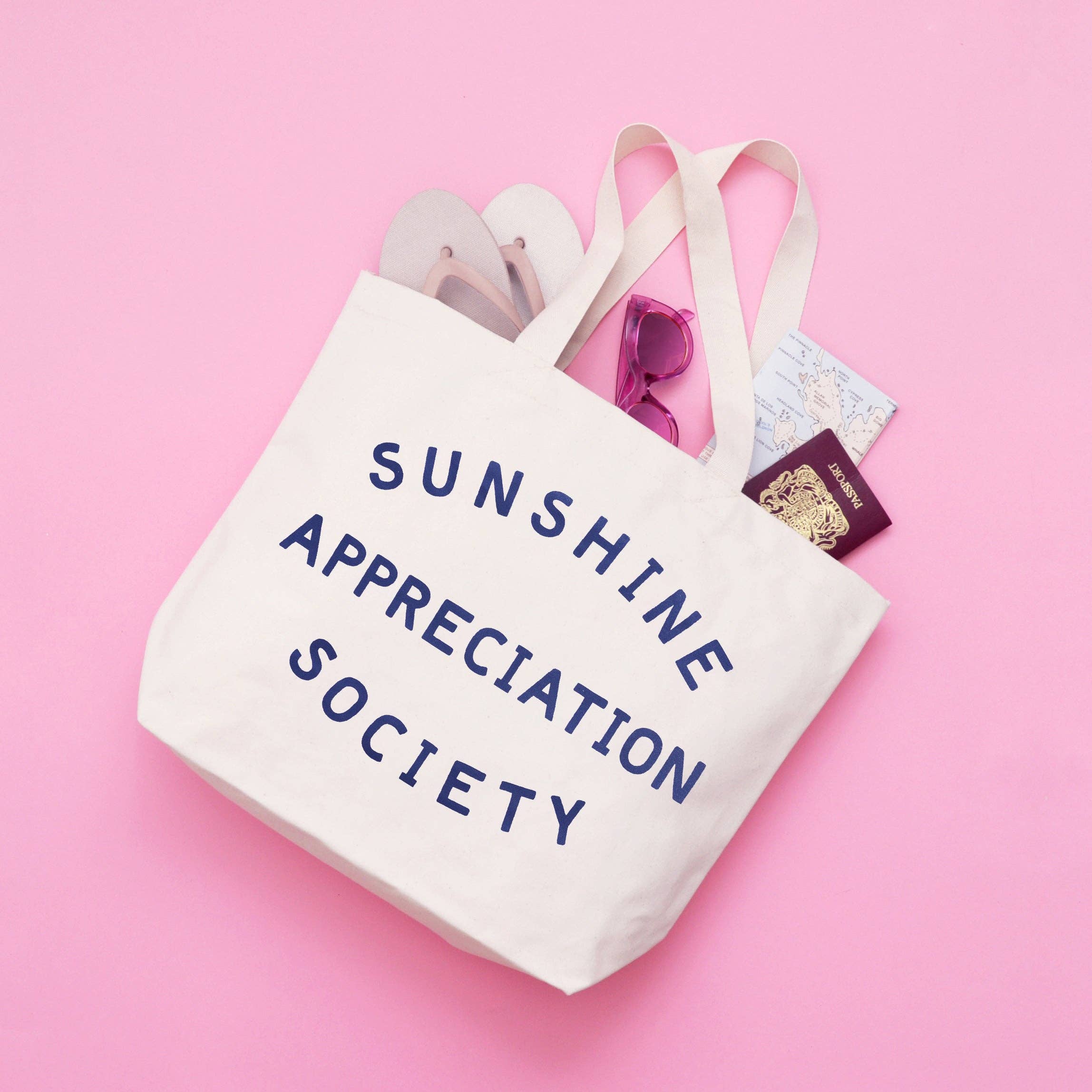 Cream canvas tote bag with “Sunshine Appreciation Society” printed in navy, filled with flip-flops, sunglasses, and travel essentials on a pink background