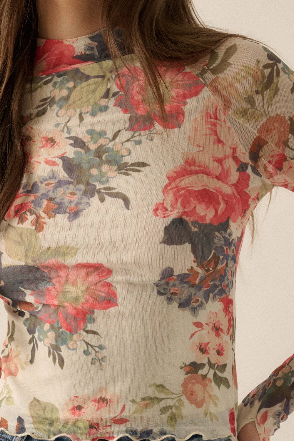 Model wearing a fitted sheer mesh long sleeve top with red, pink, and green floral print, paired with blue jeans.