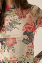 Model wearing a fitted sheer mesh long sleeve top with red, pink, and green floral print, paired with blue jeans.