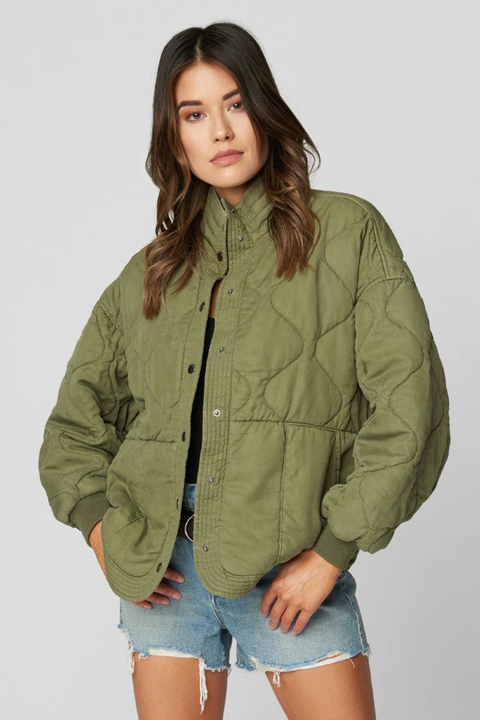 Woman wearing an olive green quilted jacket and denim shorts on a plain background