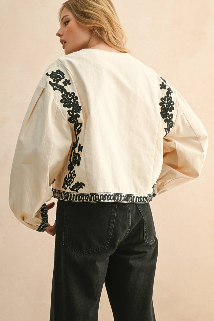 Woman wearing cream blouse with black floral embroidery, lace-up front, and long billowy sleeves.
