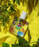 Maison Matine Nature Insolente perfume bottle with colorful leaf and beetle illustrations, placed on a vibrant yellow fabric and surrounded by green foliage.