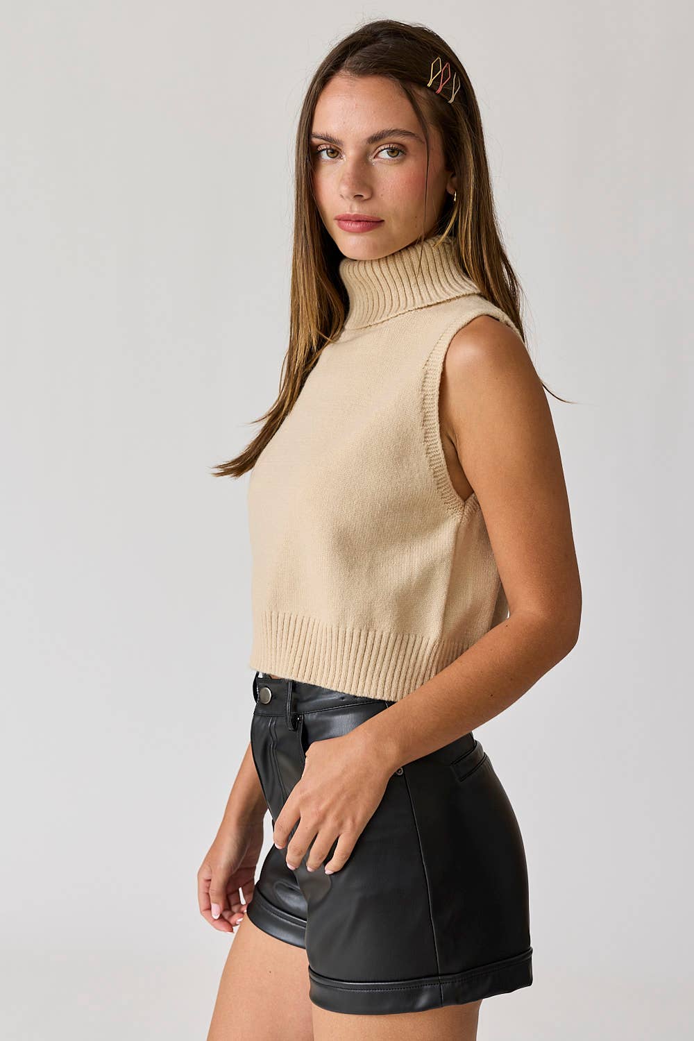 Woman wearing a beige sleeveless turtle neck knit cropped top with black leather shorts.