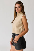 Woman wearing a beige sleeveless turtle neck knit cropped top with black leather shorts.