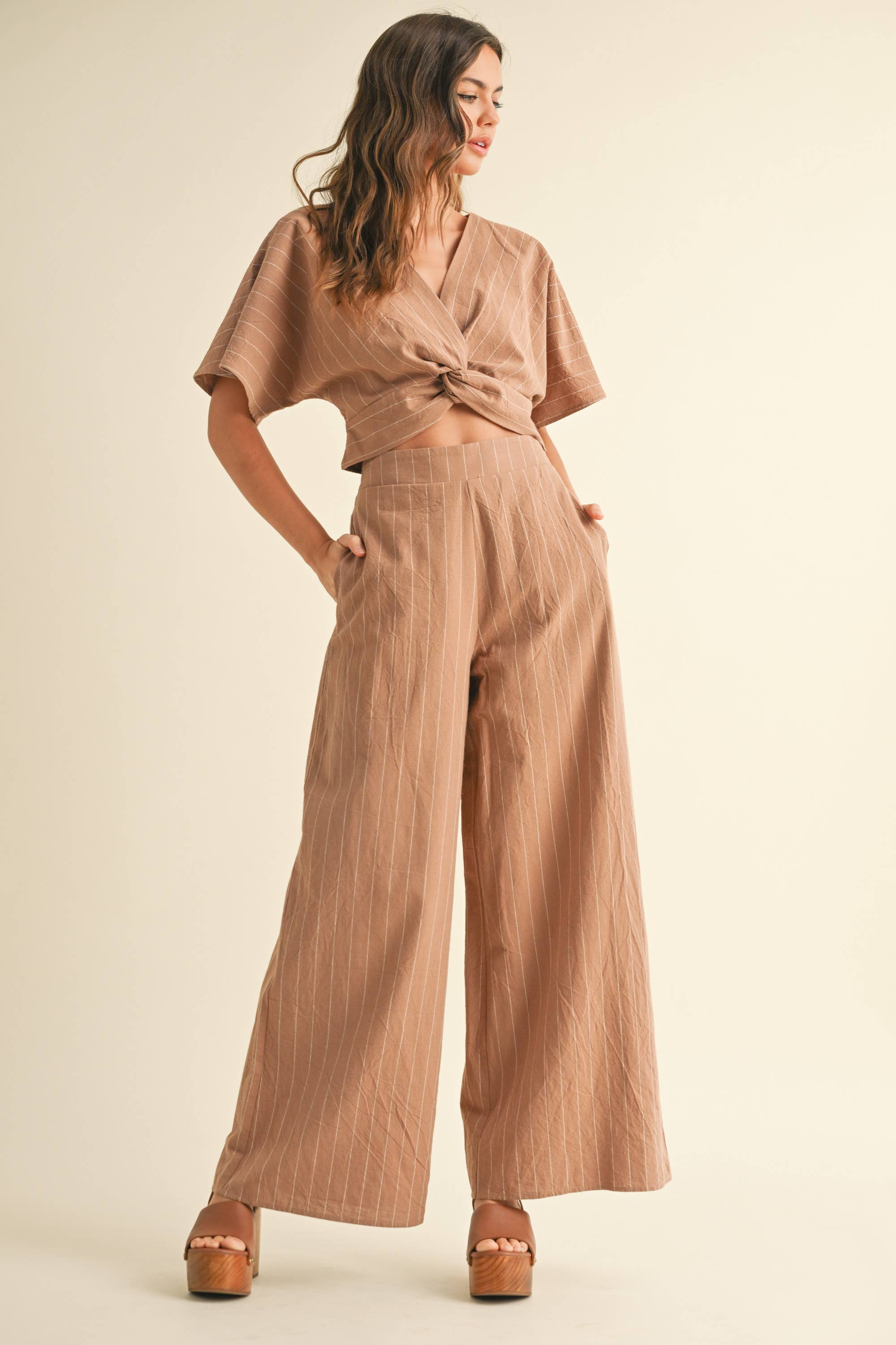 Woman wearing a two-piece striped set with a twist-front crop top and wide-leg pants in cream and tan color options.