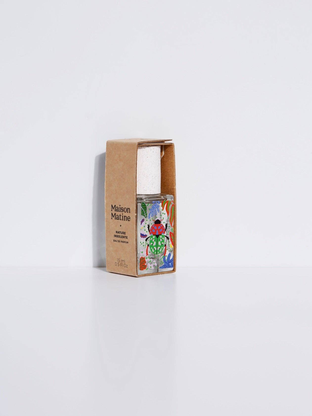 Maison Matine Nature Insolente perfume bottle with colorful leaf and beetle illustrations, placed on a vibrant yellow fabric and surrounded by green foliage.