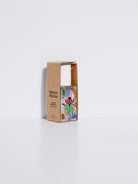 Maison Matine Nature Insolente perfume bottle with colorful leaf and beetle illustrations, placed on a vibrant yellow fabric and surrounded by green foliage.