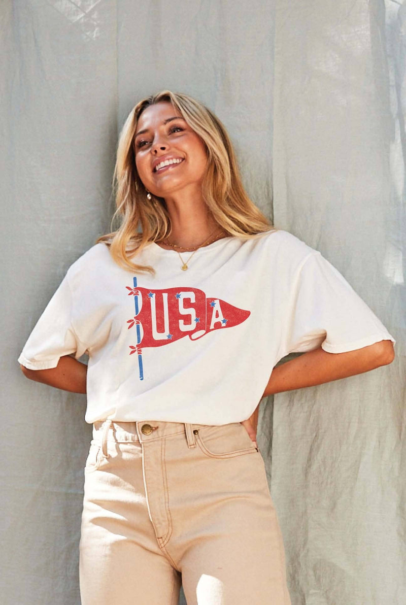 Woman wearing a cream-colored T-shirt with a vintage red USA pennant graphic, styled with beige high-waisted pants.