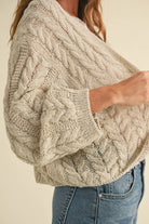 Woman wearing a loose cable knit cardigan with wide sleeves, paired with a white ribbed tank and blue denim jeans.