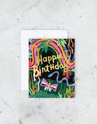 A vibrant, colorful greeting card with a hand-painted illustration that includes a butterfly and the text 'Happy Birthday'. The card is placed on a marble surface with a white envelope visible in the background.