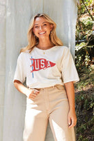 Woman wearing a cream-colored T-shirt with a vintage red USA pennant graphic, styled with beige high-waisted pants.