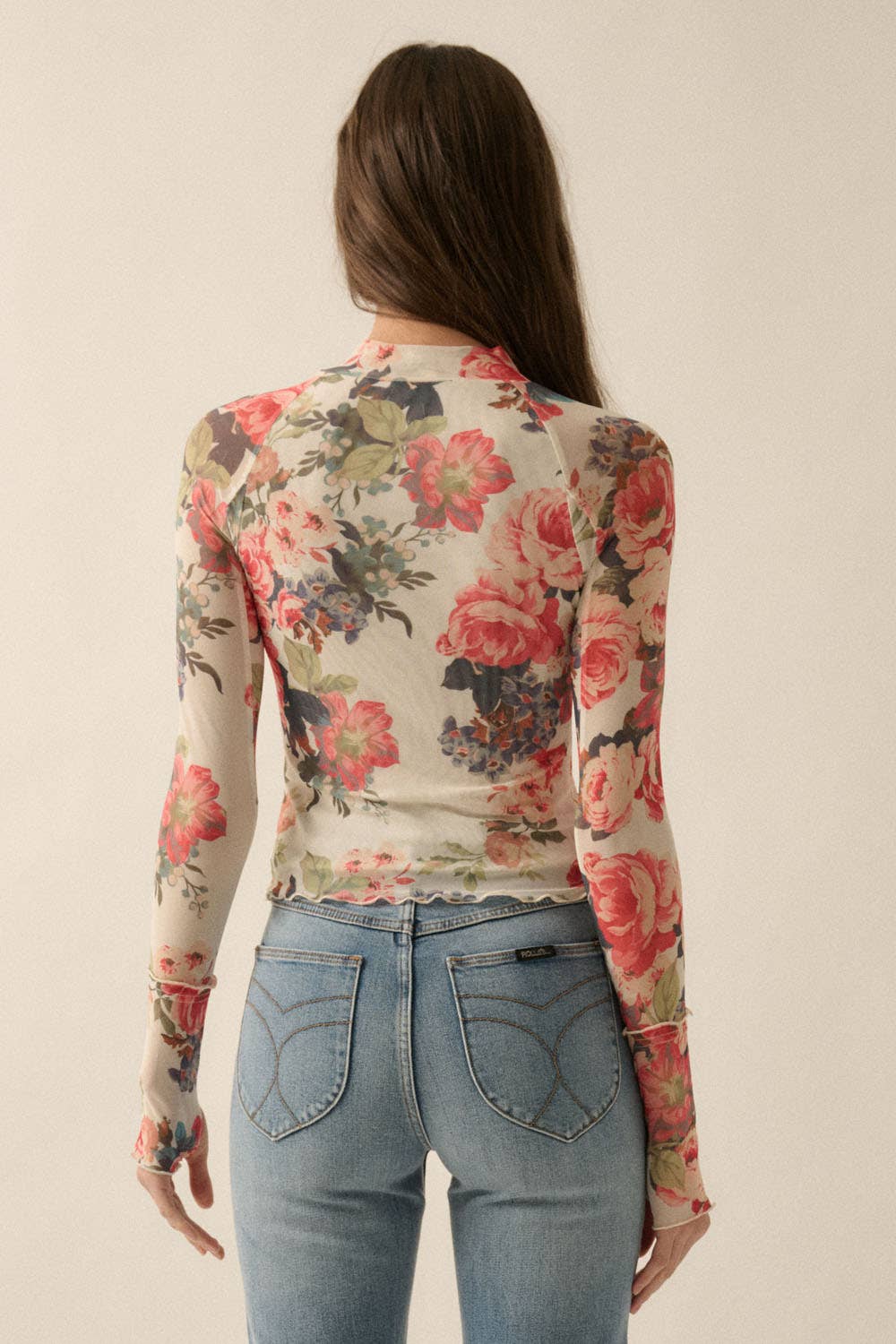 Model wearing a fitted sheer mesh long sleeve top with red, pink, and green floral print, paired with blue jeans.