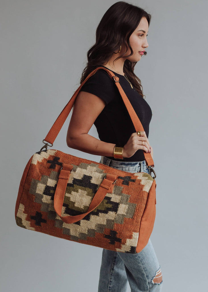 Earth-tone woven weekender bag with geometric pattern in black, green, and cream, held by woman in jeans and black top