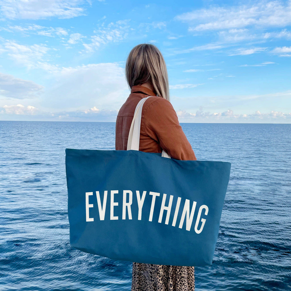 Oversized navy blue tote bag with cream handles and bold white “EVERYTHING” text, held by a hand against a light wood background