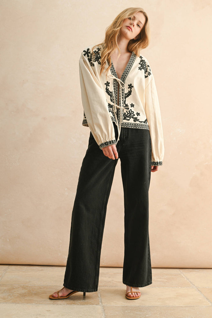 Woman wearing cream blouse with black floral embroidery, lace-up front, and long billowy sleeves.