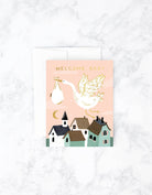 Greeting card with a stork carrying a baby bundle flying above pastel houses, with “Welcome Baby” text.