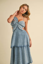 Woman wearing a denim maxi dress with a large front bow, side cutouts, spaghetti straps, and tiered skirt.