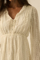 Woman wearing a cream mini dress with sheer long sleeves, button front, and tie details.