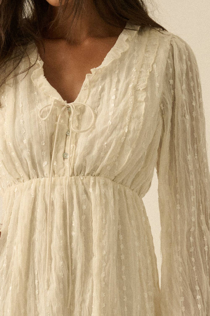 Woman wearing a cream mini dress with sheer long sleeves, button front, and tie details.