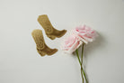 A brass bookmark shaped like a cowgirl boot with floral engravings, displayed on a daisy-patterned yellow backing card.