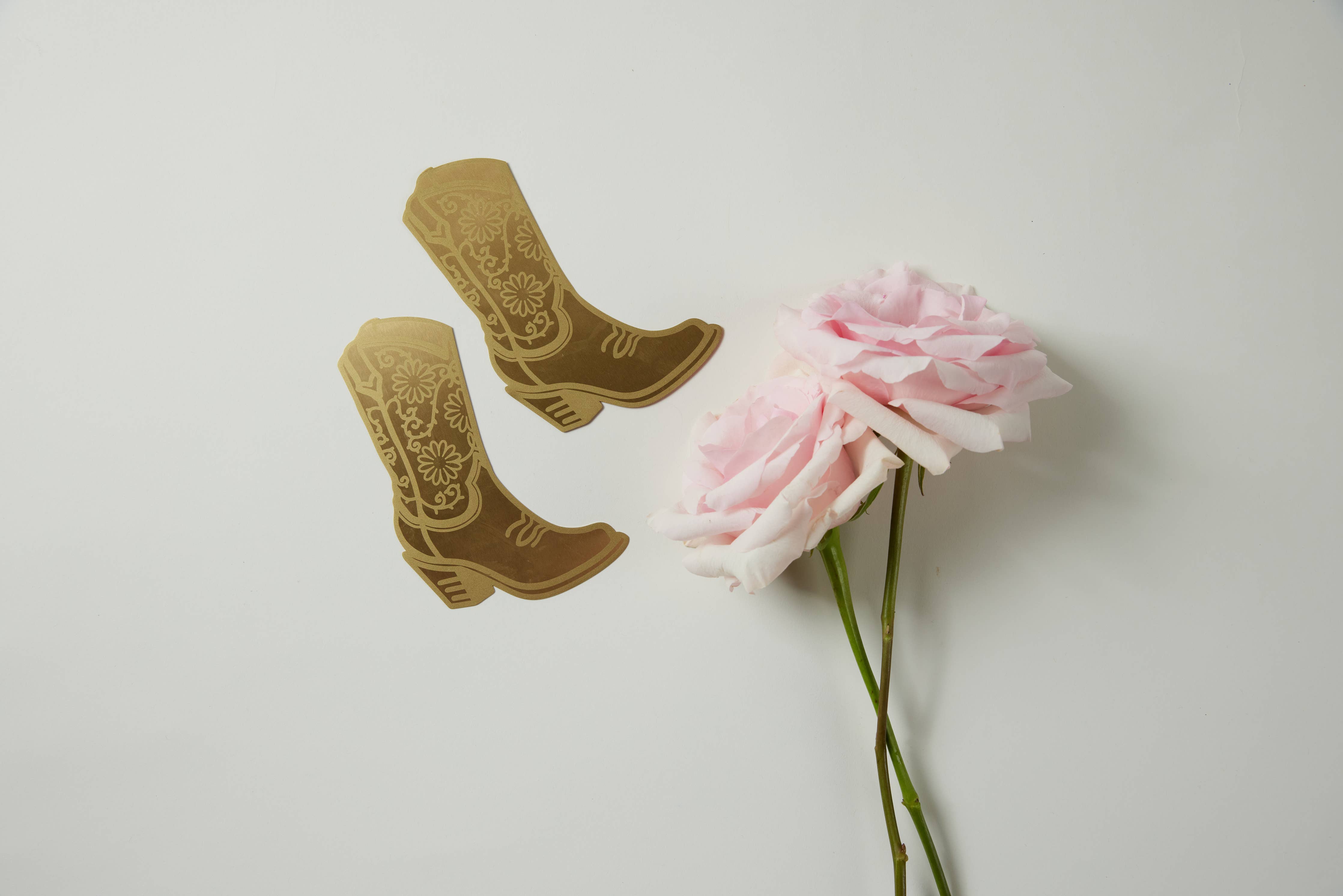 A brass bookmark shaped like a cowgirl boot with floral engravings, displayed on a daisy-patterned yellow backing card.