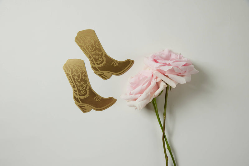 A brass bookmark shaped like a cowgirl boot with floral engravings, displayed on a daisy-patterned yellow backing card.