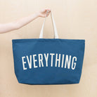 Oversized navy blue tote bag with cream handles and bold white “EVERYTHING” text, held by a hand against a light wood background