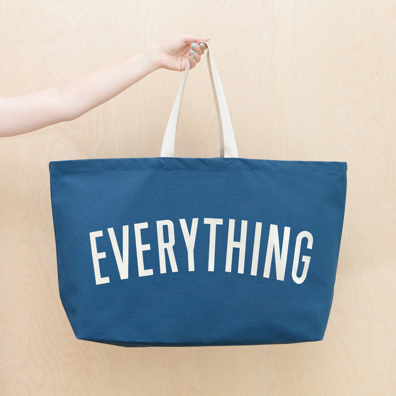 Oversized navy blue tote bag with cream handles and bold white “EVERYTHING” text, held by a hand against a light wood background
