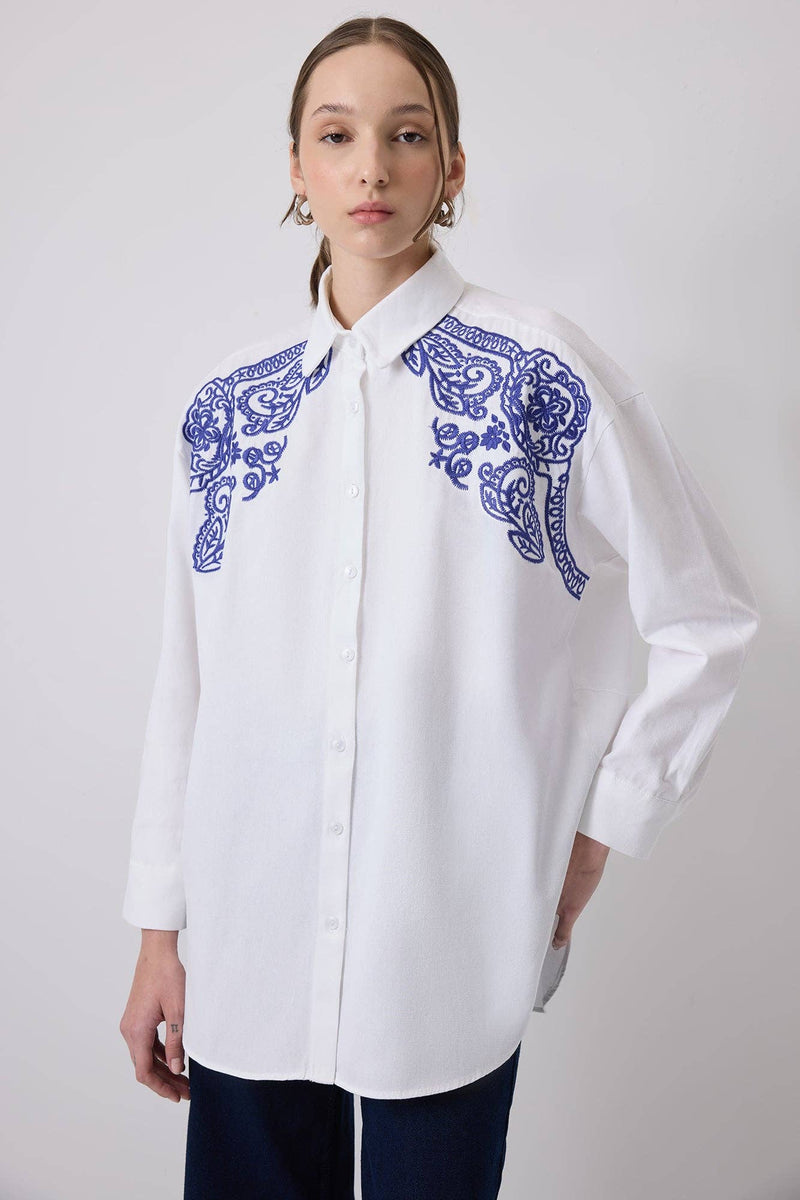 Woman wearing a white oversized shirt with bold blue embroidery on shoulders