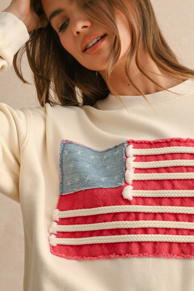 Woman wearing a cream sweater with a textured rope-stripe American flag design, paired with light wash jeans.