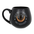 Black mug with ‘Let Me Consult My Crystals’ text and a gold foil crescent moon and crystal design, placed among crystals.
