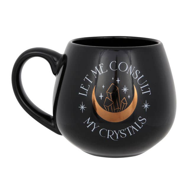 Black mug with ‘Let Me Consult My Crystals’ text and a gold foil crescent moon and crystal design, placed among crystals.