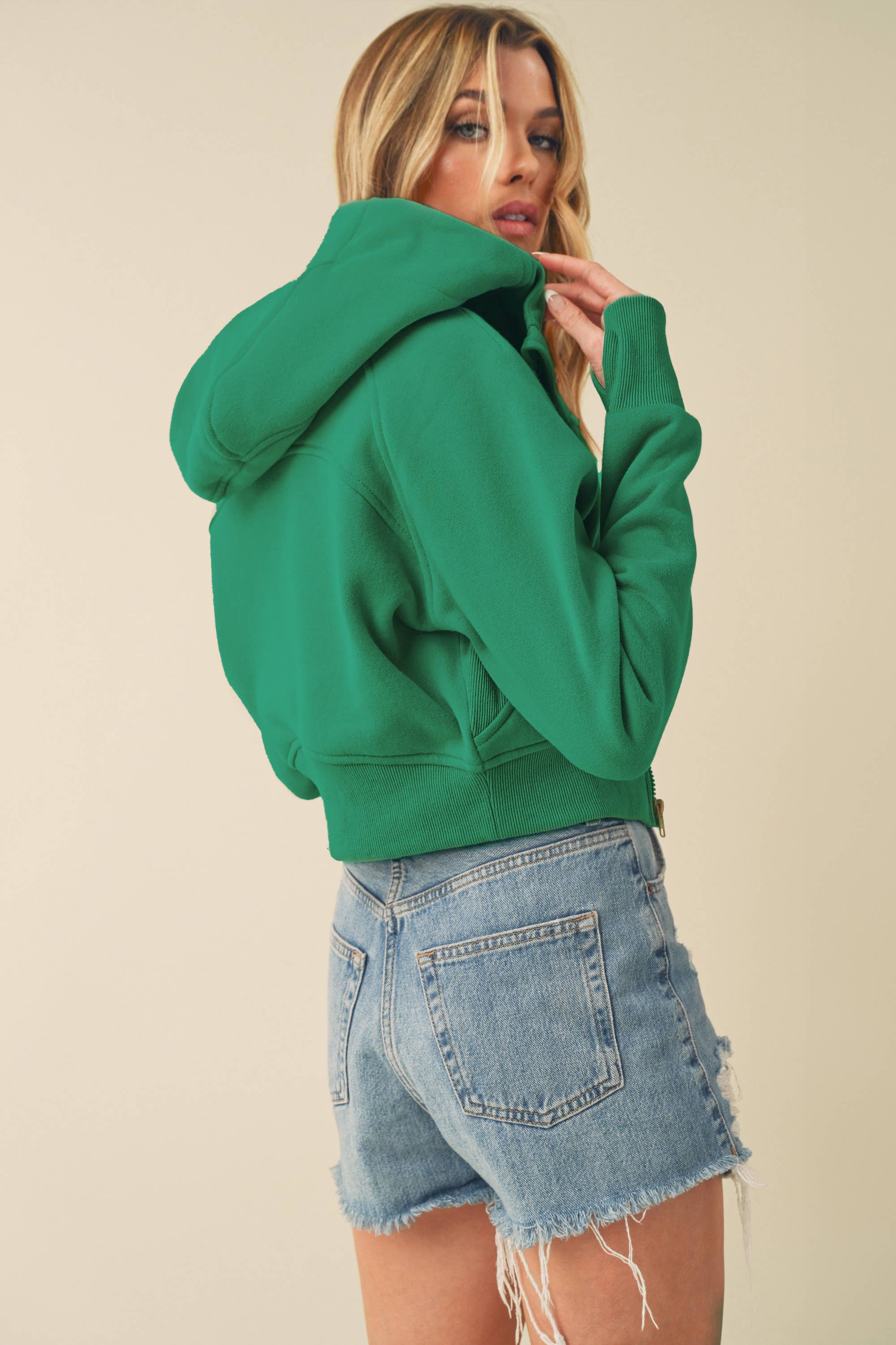 Model wearing a cropped zip-up hoodie with adjustable hood and ribbed cuffs, paired with distressed denim shorts.