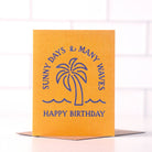 Orange birthday card with blue text saying ‘Sunny Days & Many Waves, Happy Birthday,’ featuring a palm tree illustration.