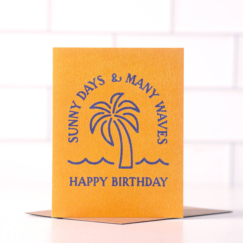 Orange birthday card with blue text saying ‘Sunny Days & Many Waves, Happy Birthday,’ featuring a palm tree illustration.