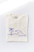 Folded cream-colored t-shirt with a blue minimalist palm tree, wave, and "LIVE LOVE SURF" graphic.