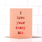 A blush-colored greeting card with "I love your crazy ass" in red and orange watercolor-style text.