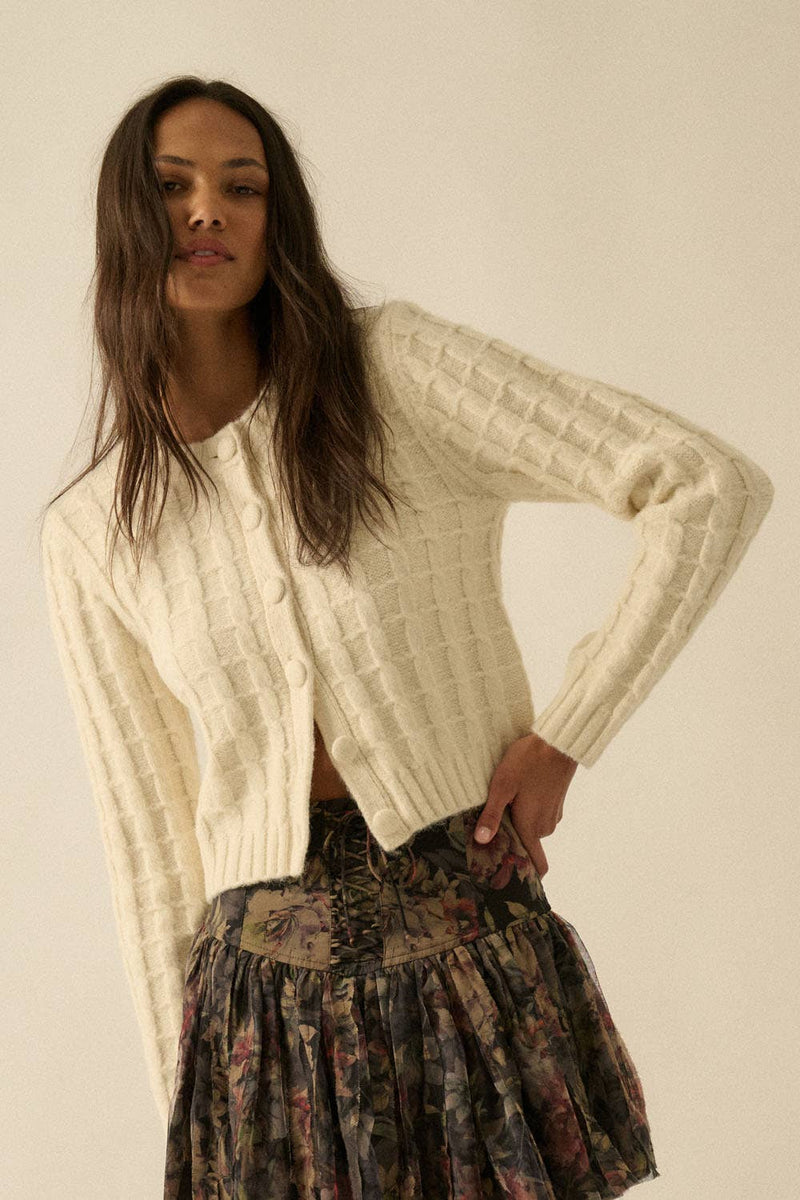 Woman wearing a cream textured knit cardigan with covered buttons, styled with a floral skirt.