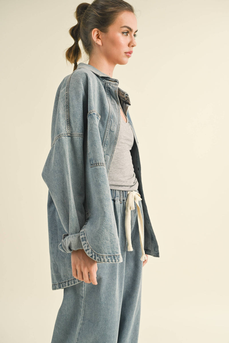 Model in an oversized denim jacket with front pockets. First image: jacket buttoned, paired with white shorts. Second image: jacket open, over a grey tank and denim pants.