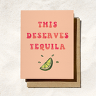 A peach-colored greeting card with red text reading "This Deserves Tequila" and a lime slice illustration, placed on a brown envelope.