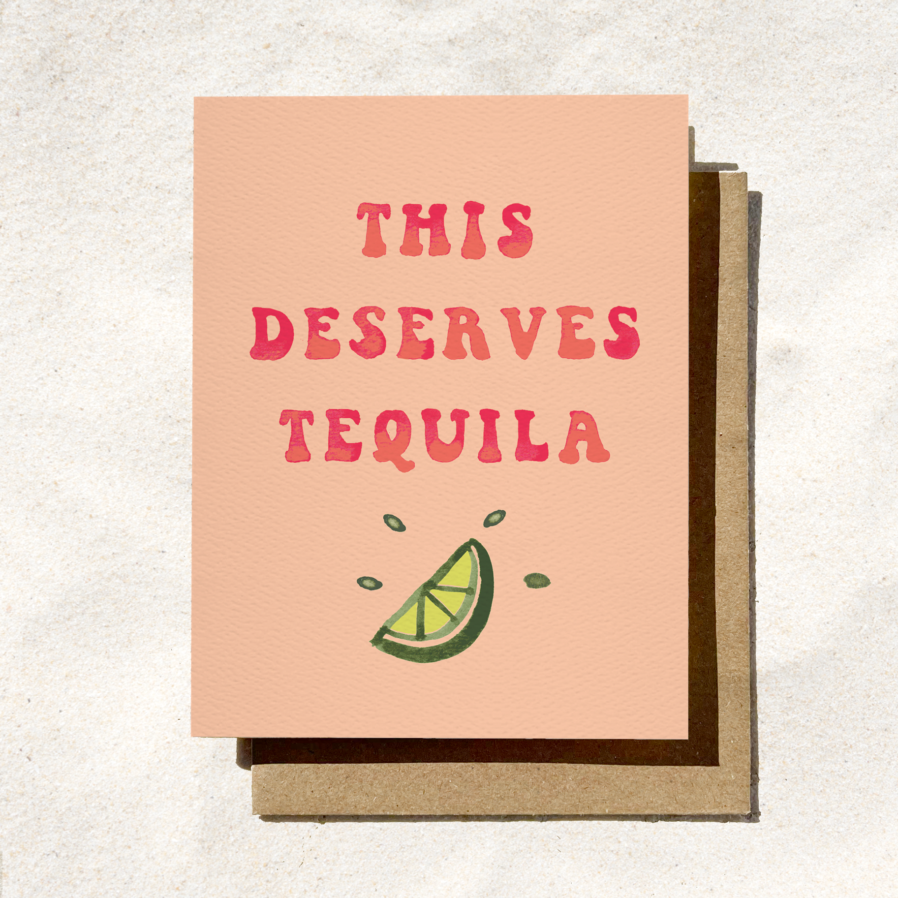 A peach-colored greeting card with red text reading "This Deserves Tequila" and a lime slice illustration, placed on a brown envelope.